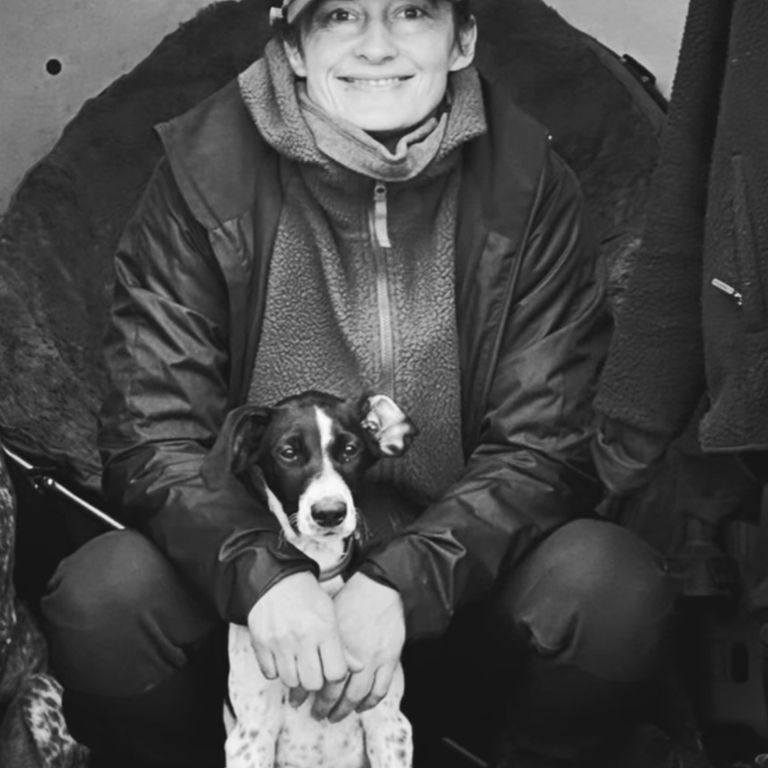 Lächelnde Person mit Hund in Outdoor-Kleidung und Basecap sitzt mit treuem Vierbeiner.