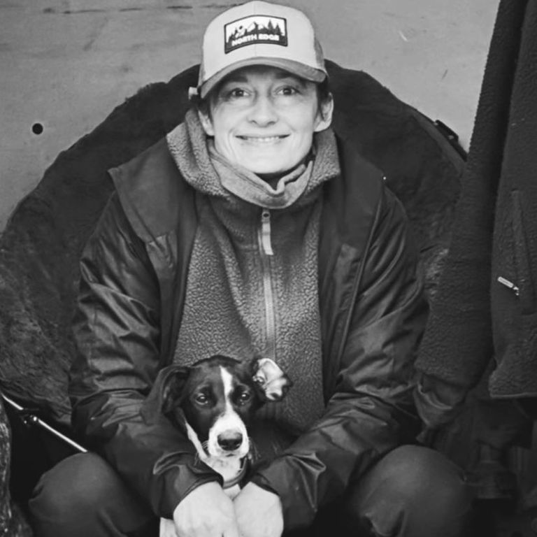Lächelnde Person mit Hund in Outdoor-Kleidung und Basecap sitzt mit treuem Vierbeiner.
